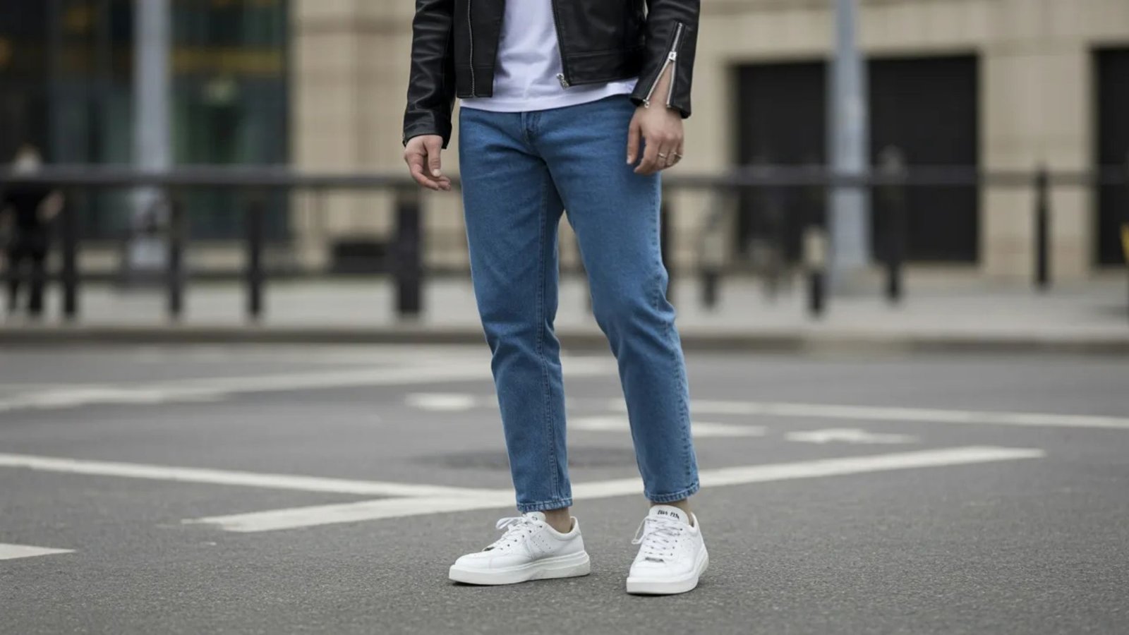 A man wearing blue jeans and a black leather jacket stands confidently, showcasing a casual yet stylish look.