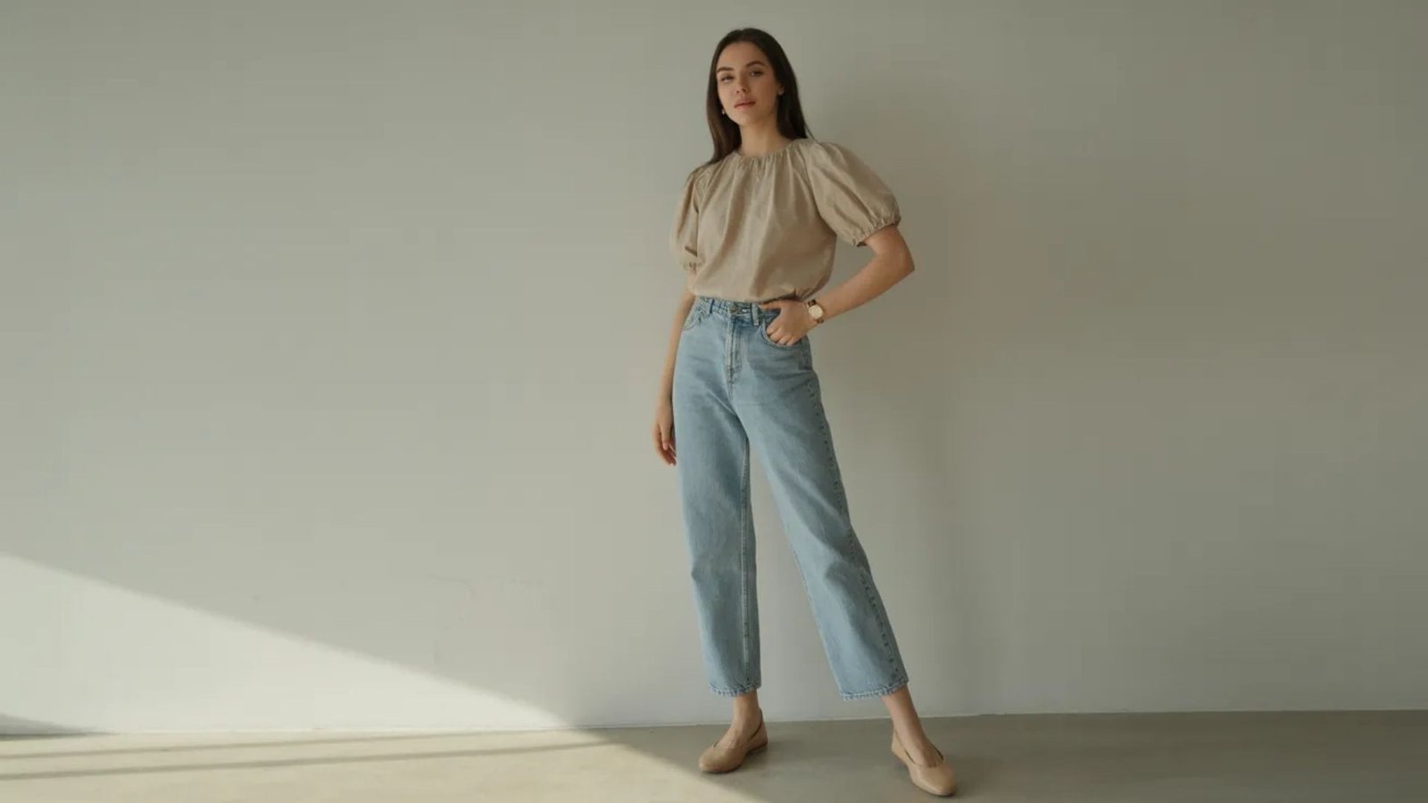 A woman in a beige blouse and jeans stands confidently, showcasing a casual yet stylish outfit.