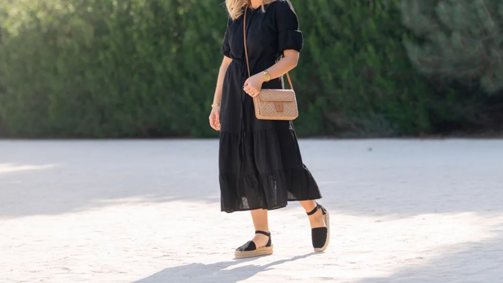 A woman wearing a black dress and sandals strolls on the beach, with the ocean waves in the background.