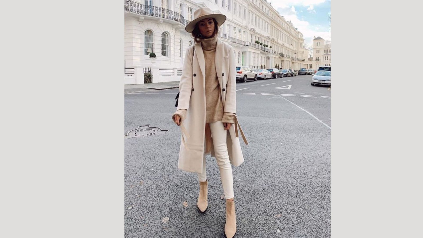 A woman in a white coat and hat stands on the street, looking confidently ahead.