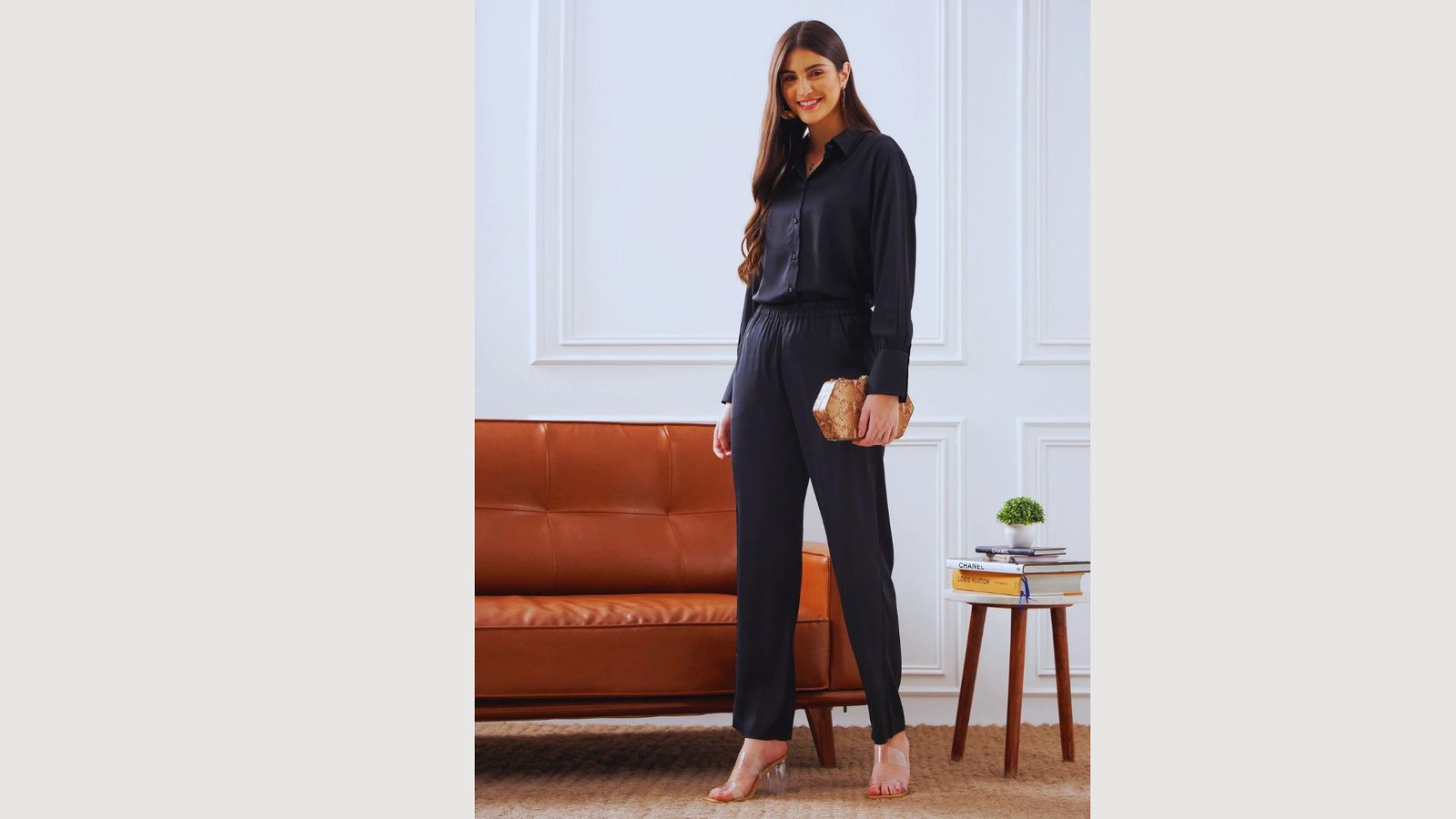 A woman in a black jumpsuit stands confidently on a couch, showcasing a stylish and bold fashion choice.
