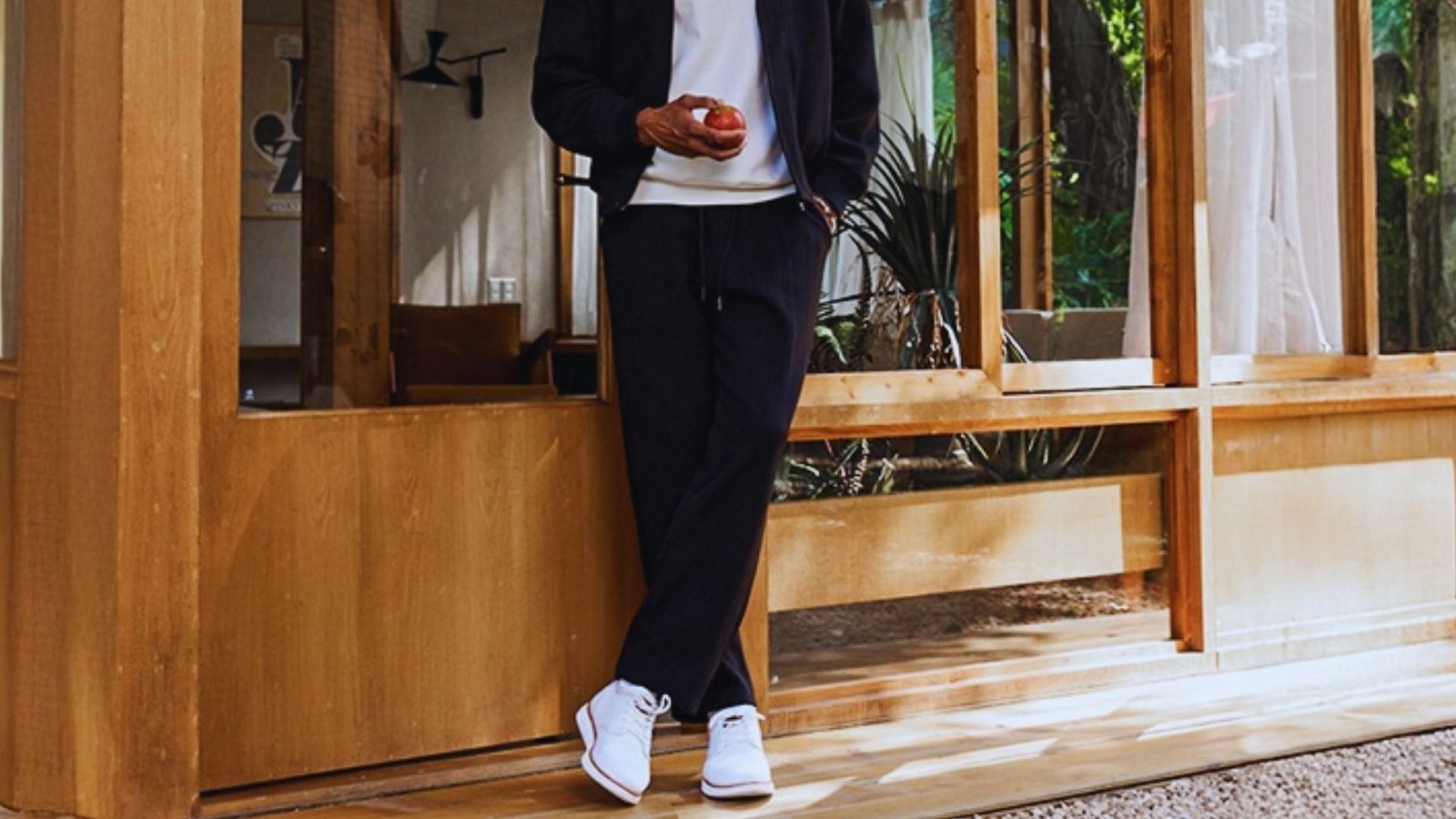 A man in a black jacket and white sneakers leans against a wooden wall, looking relaxed and casual.