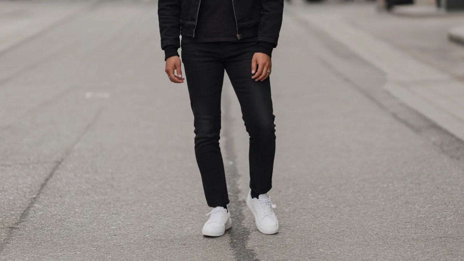 A man wearing a black jacket and jeans stands on a street, looking confidently ahead.