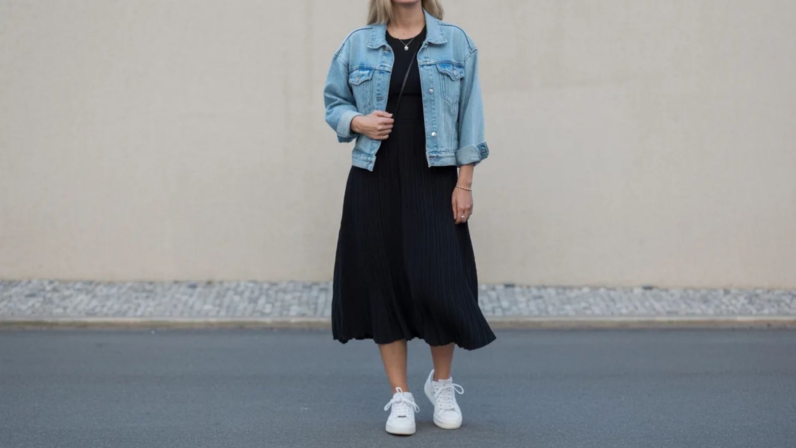 A woman wearing a black dress paired with a denim jacket stands confidently, showcasing a stylish casual look.