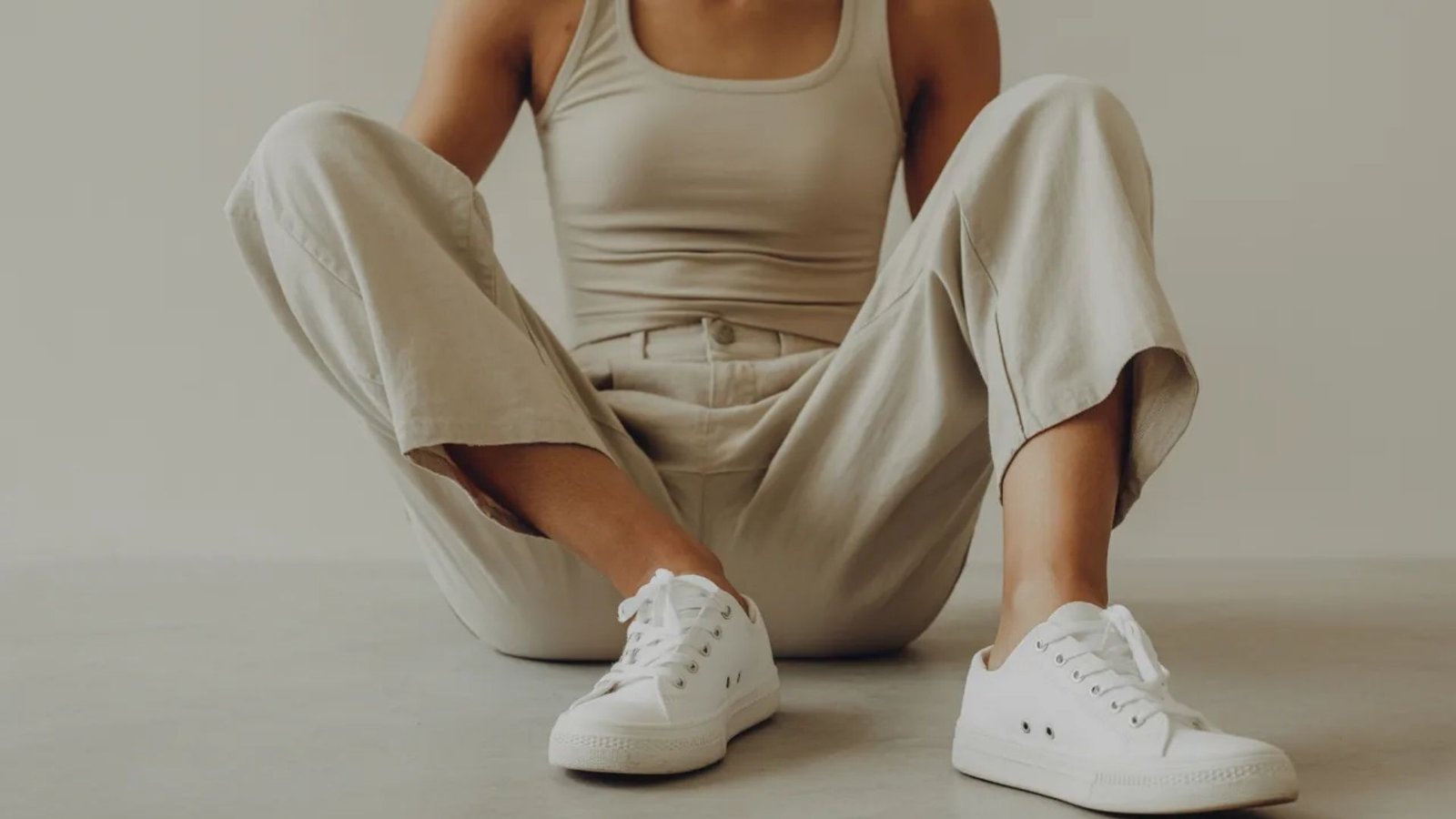 A woman wearing a tank top and pants is sitting on the floor, looking relaxed and contemplative.