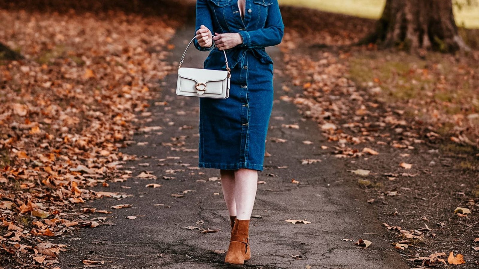 A woman in a denim dress and boots walks along a scenic path surrounded by greenery.