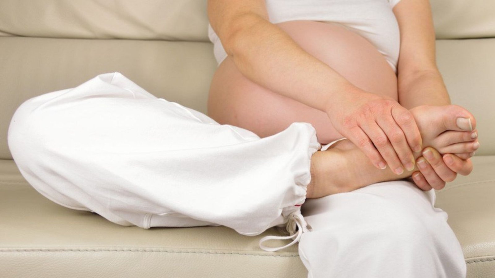 A pregnant woman sits on a couch, gently resting her hands on her stomach, looking serene and contemplative.