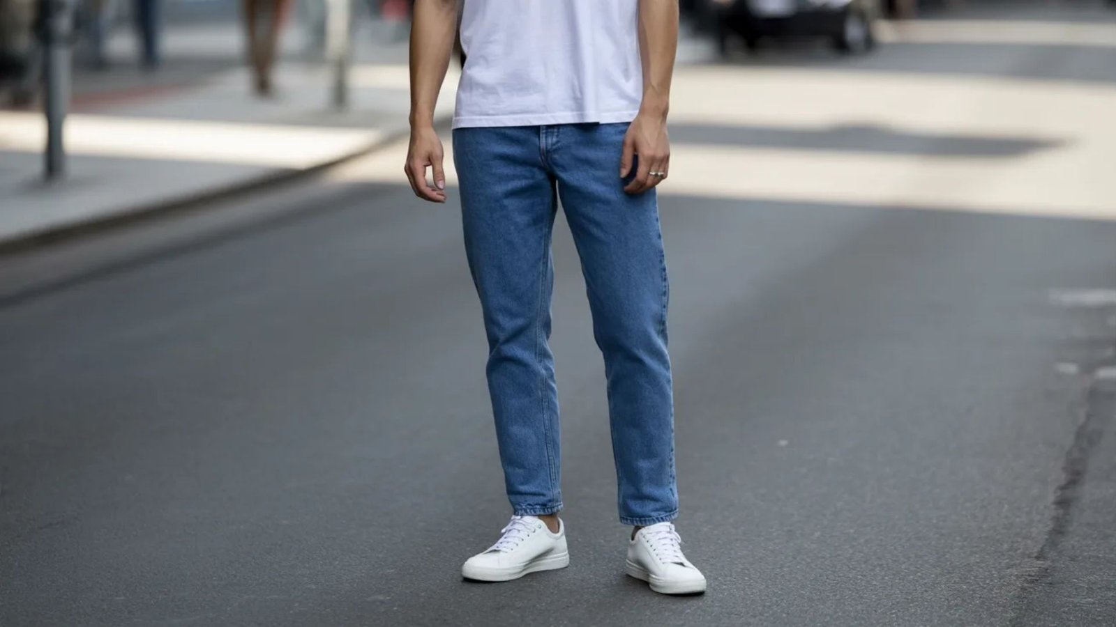 A man wearing a white t-shirt and blue jeans stands on a street, looking towards the camera.
