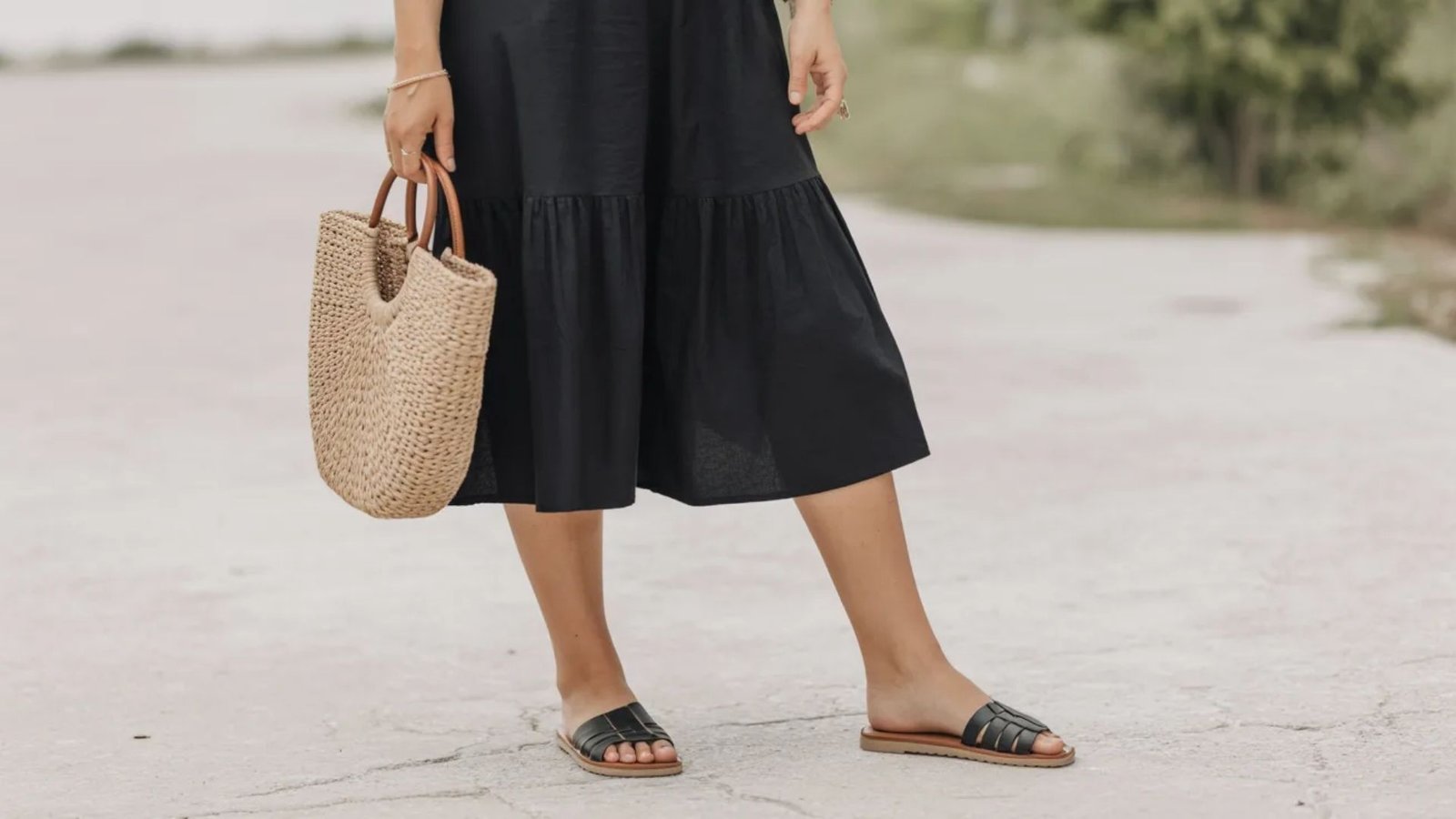 A woman in a black dress and straw sandals stands confidently, showcasing a stylish summer outfit.