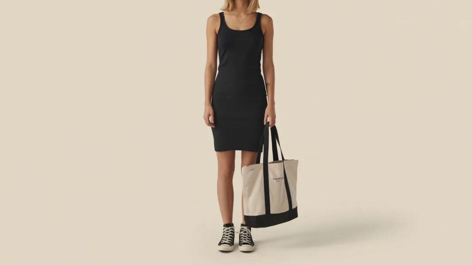 A woman in a black dress and sneakers holds a tote bag, standing confidently with a casual yet stylish look.