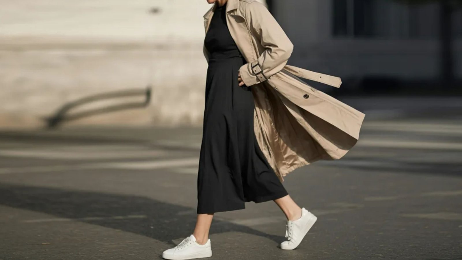 A woman in a black dress and trench coat walks confidently down the street, surrounded by urban scenery.