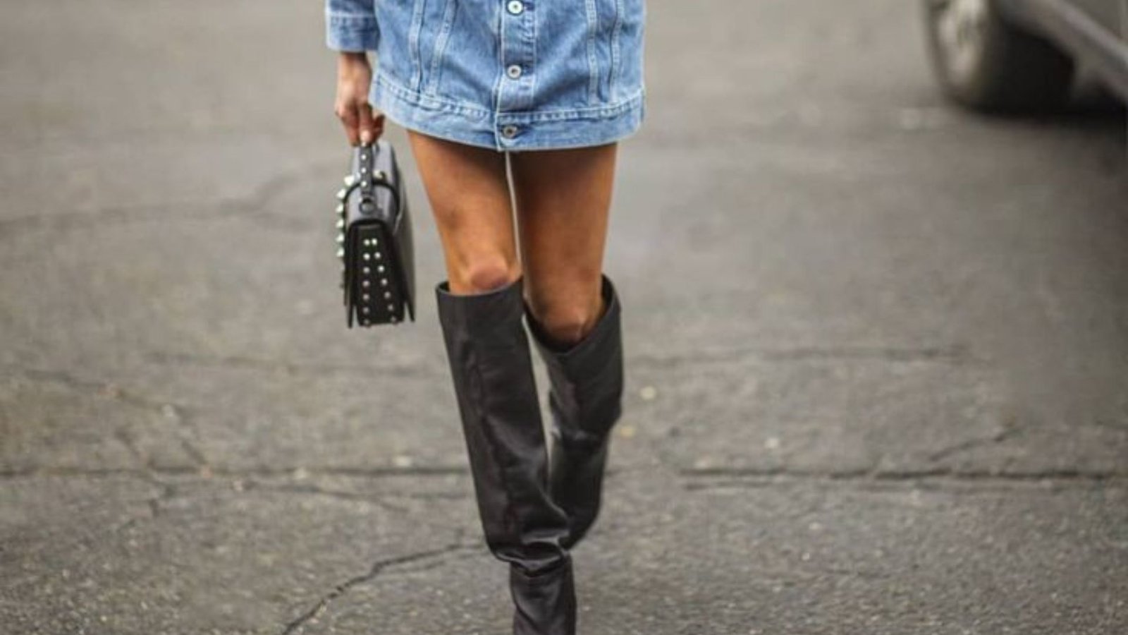 A woman in a denim dress and boots strolls down a city street, surrounded by urban scenery.
