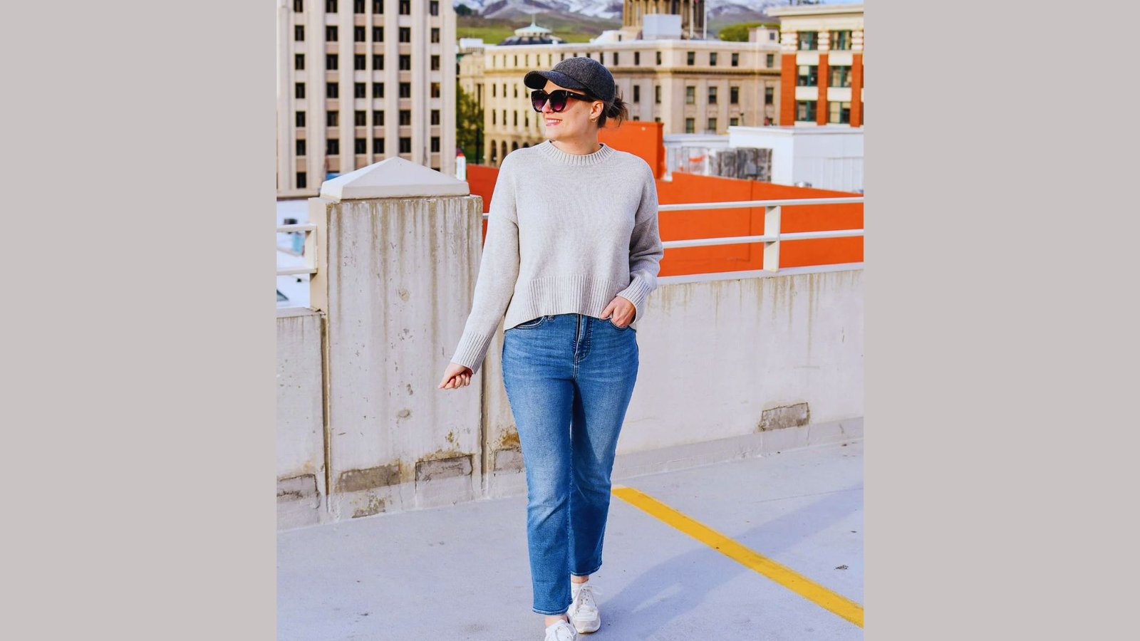A woman in jeans and a sweater stands confidently on a rooftop, overlooking the city skyline.