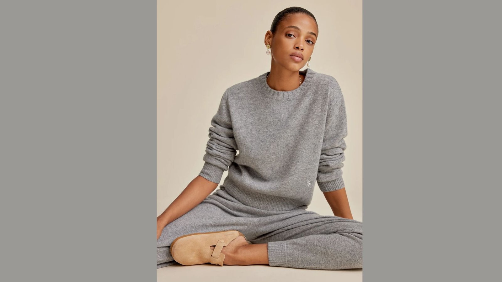 A woman in a grey sweater and pants, sitting cross-legged on the floor, appearing thoughtful and at ease.