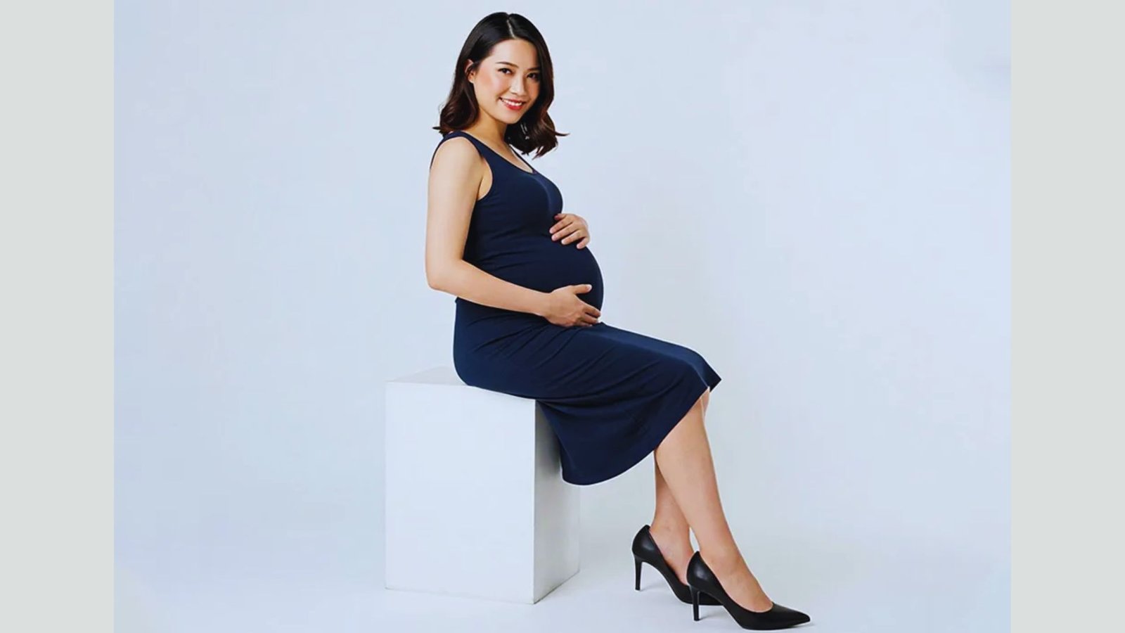A pregnant woman in a blue dress sits on a white box, smiling gently while resting her hands on her belly.