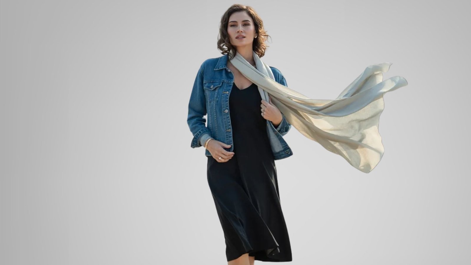 A woman in a black dress and denim jacket holds a white scarf, standing confidently with a casual yet stylish look.