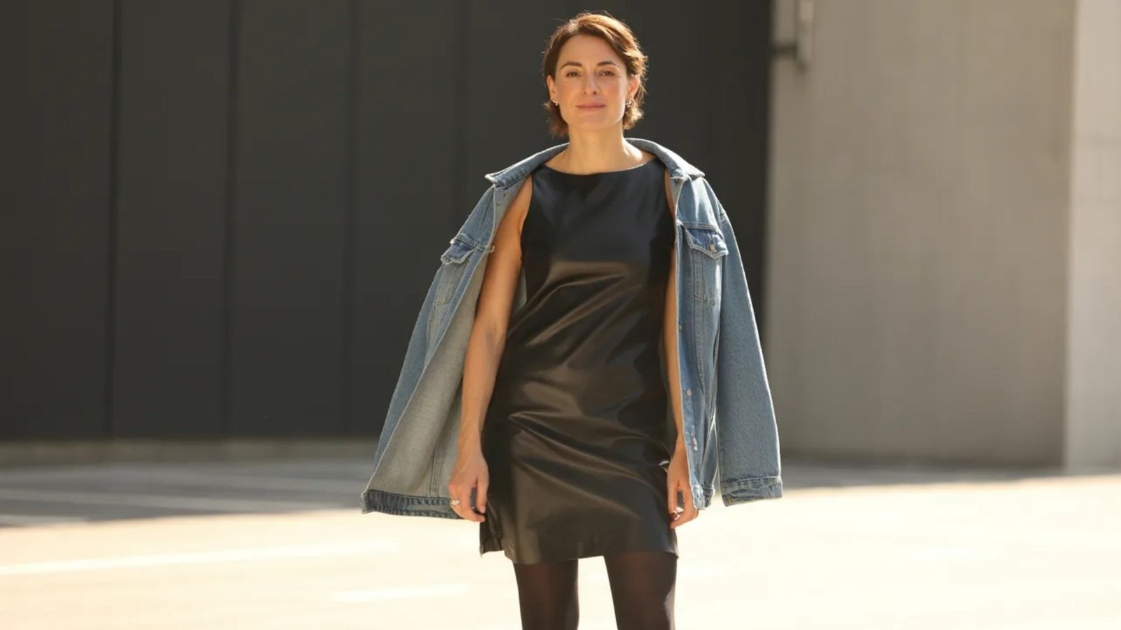 A woman wearing a black leather dress paired with a denim jacket stands confidently, showcasing her stylish outfit.