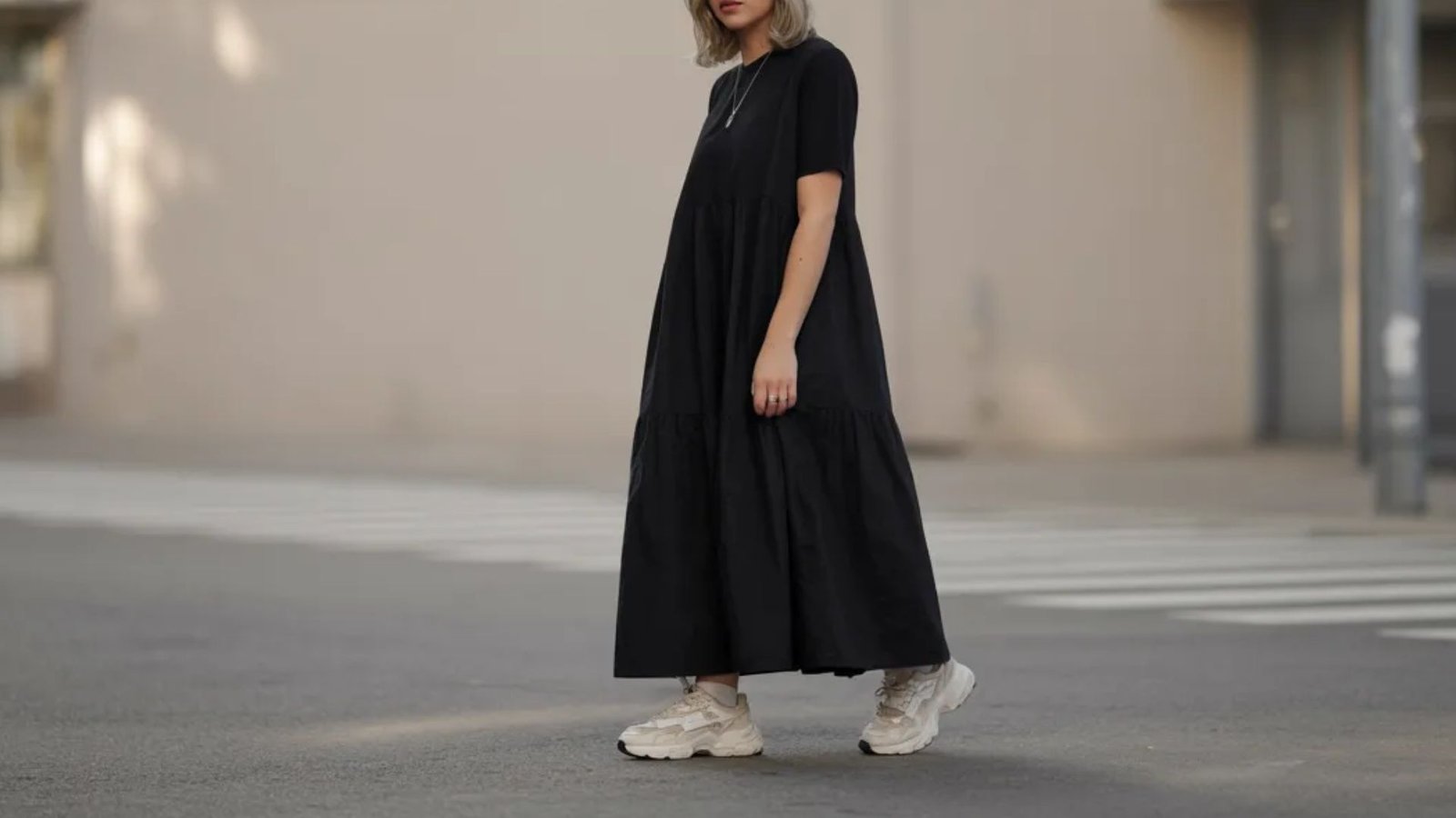 woman in a stylish black dress and sneakers stands on the sidewalk, surrounded by urban scenery.