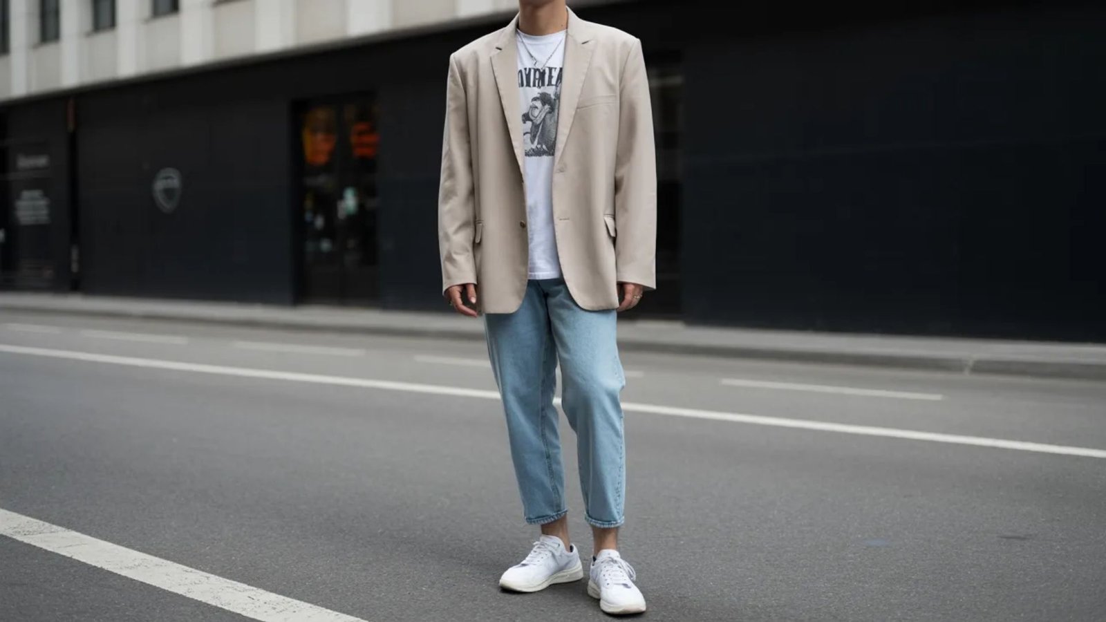 A man dressed in a beige blazer and jeans stands on the sidewalk in an urban setting.