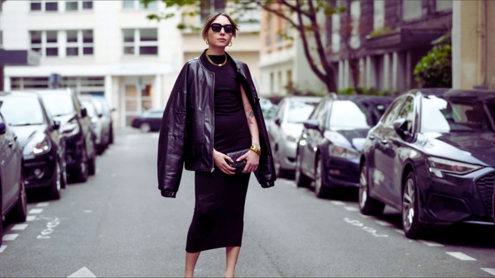 A woman in a black sheath dress and sunglasses stands confidently on a city street.