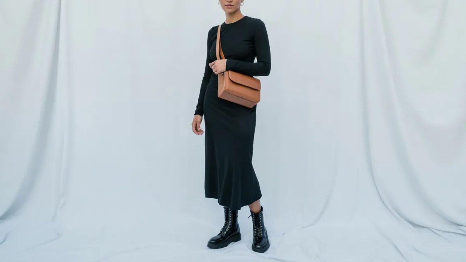 A woman wearing a black dress and boots carries a brown bag, showcasing a stylish and modern look.