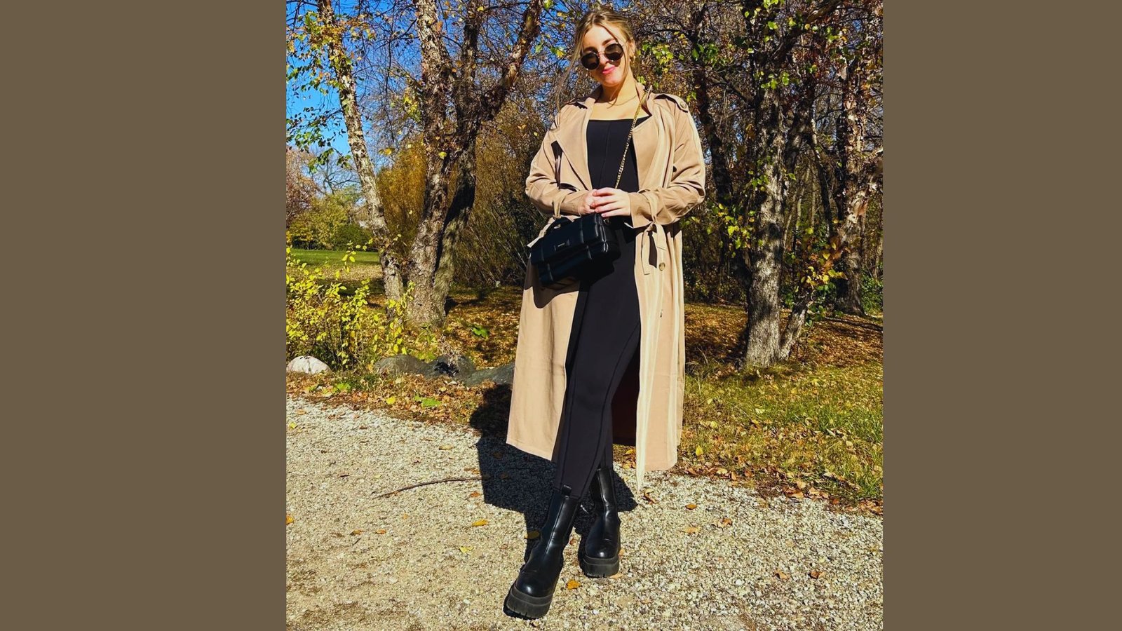  A woman wearing a black dress and long trench coat stands on a road, exuding elegance and poise.
