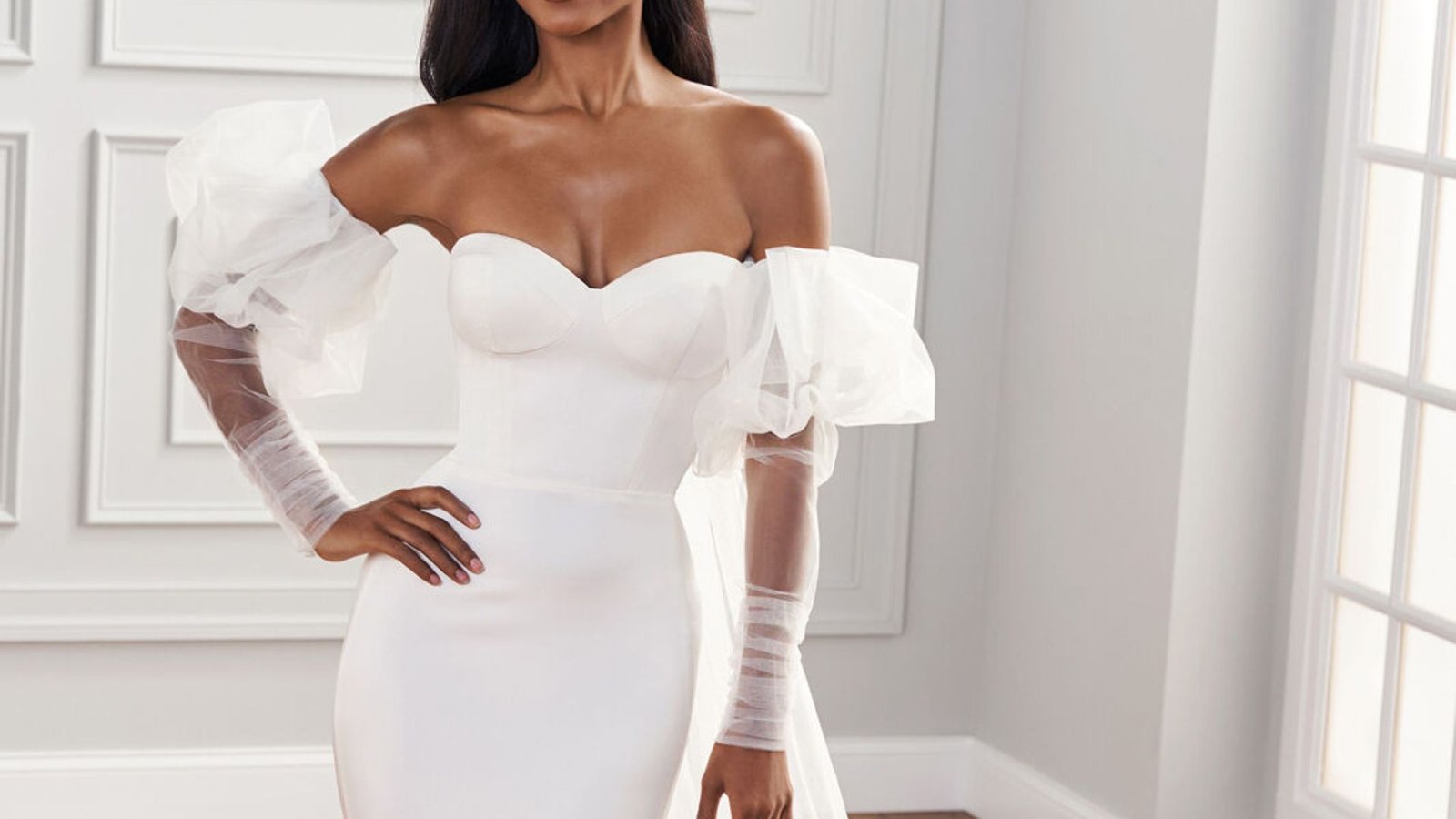 A model poses for the camera in an elegant white wedding dress, showcasing intricate lace and a flowing silhouette.