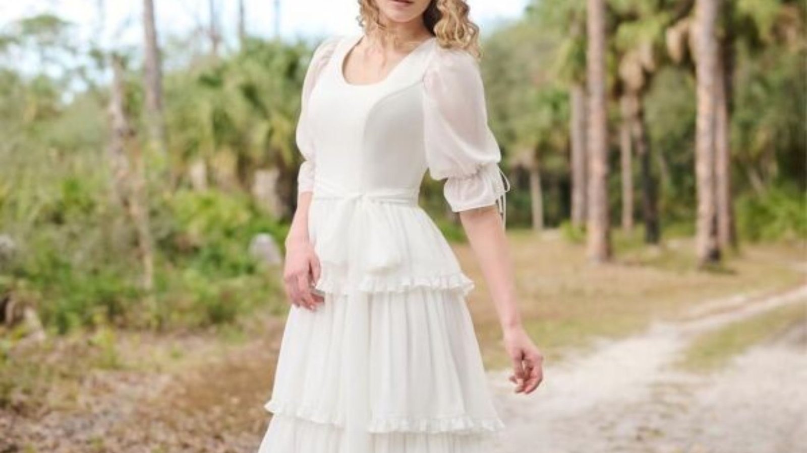 A woman in a white dress stands on a dirt road, surrounded by greenery under a clear blue sky.