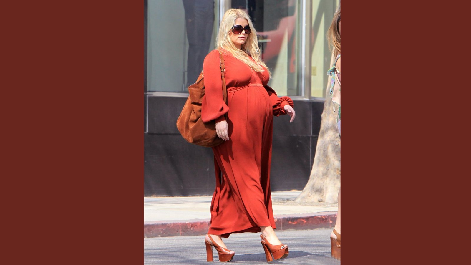 A pregnant woman in a red dress strolls down the street, showcasing her baby bump and enjoying the day.