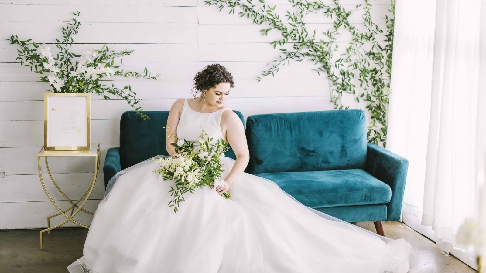 A bride in a white gown sits gracefully on a blue couch in a bright, white room.