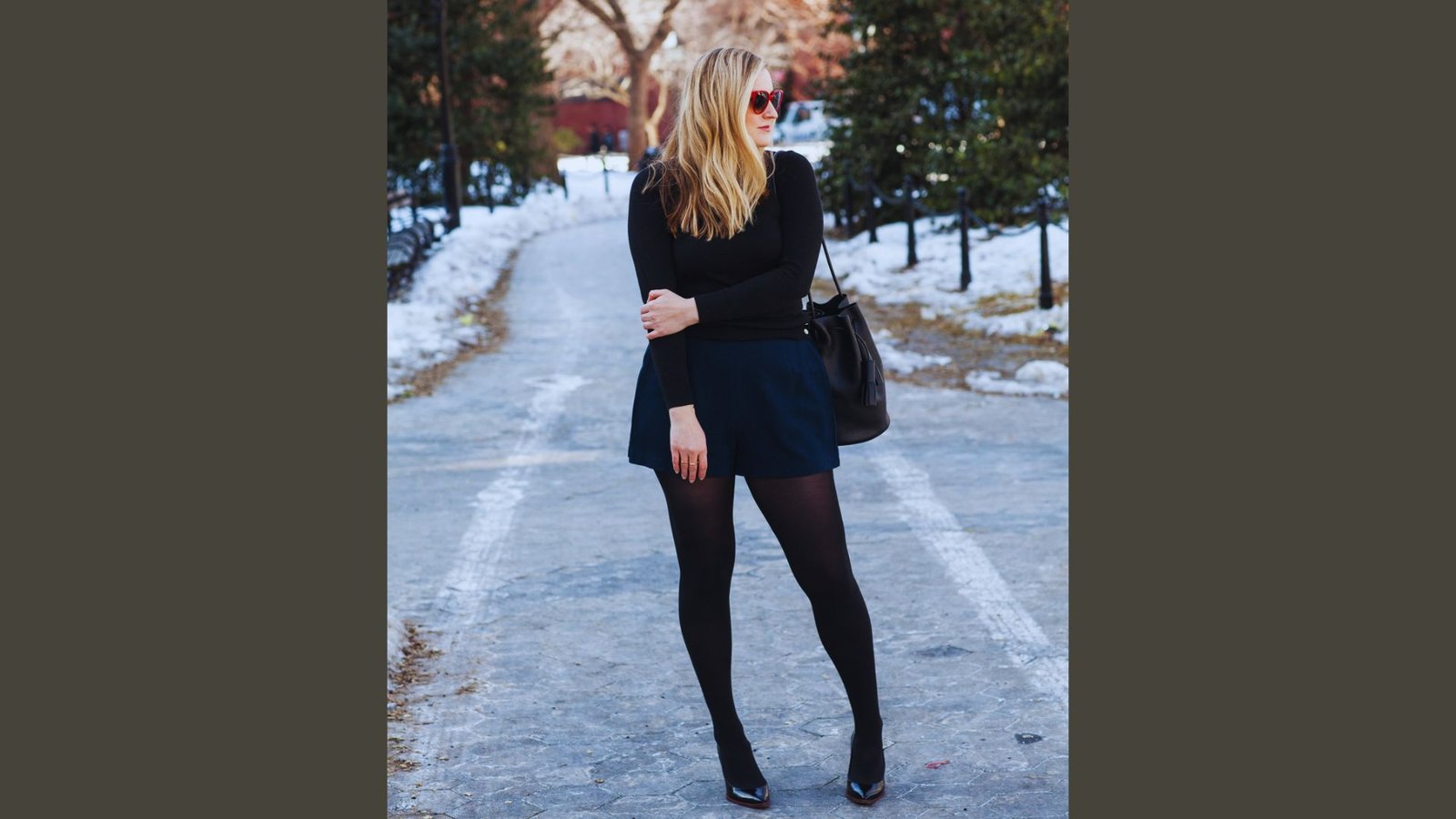 A woman in a black sweater and skirt stands on a snowy street, surrounded by a winter landscape.