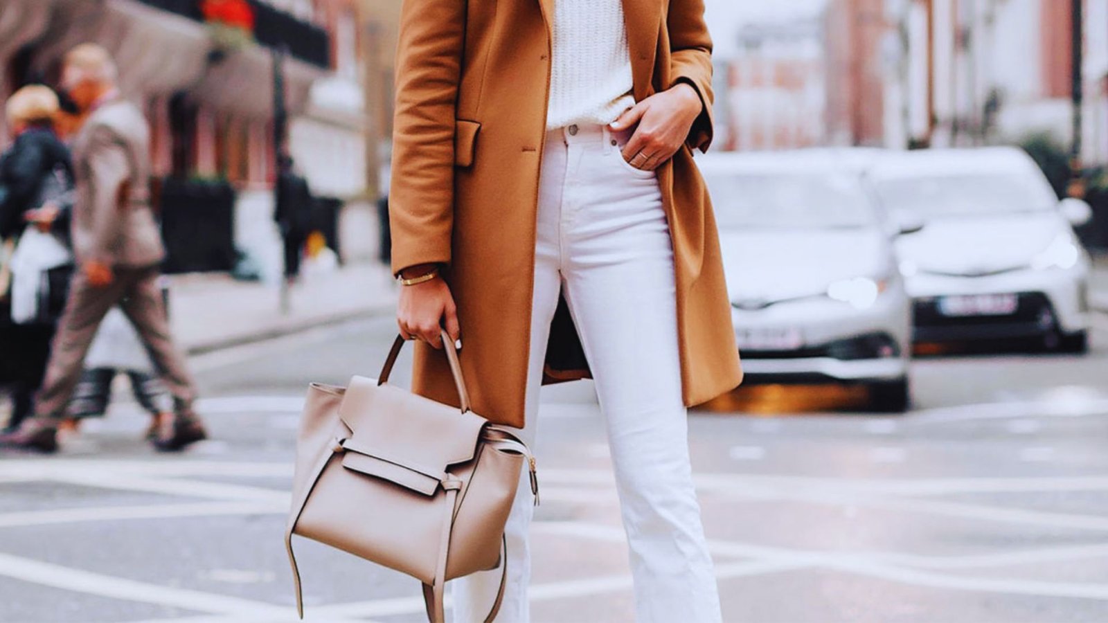 A woman wearing white jeans and a camel coat stands confidently, showcasing a stylish and modern outfit.