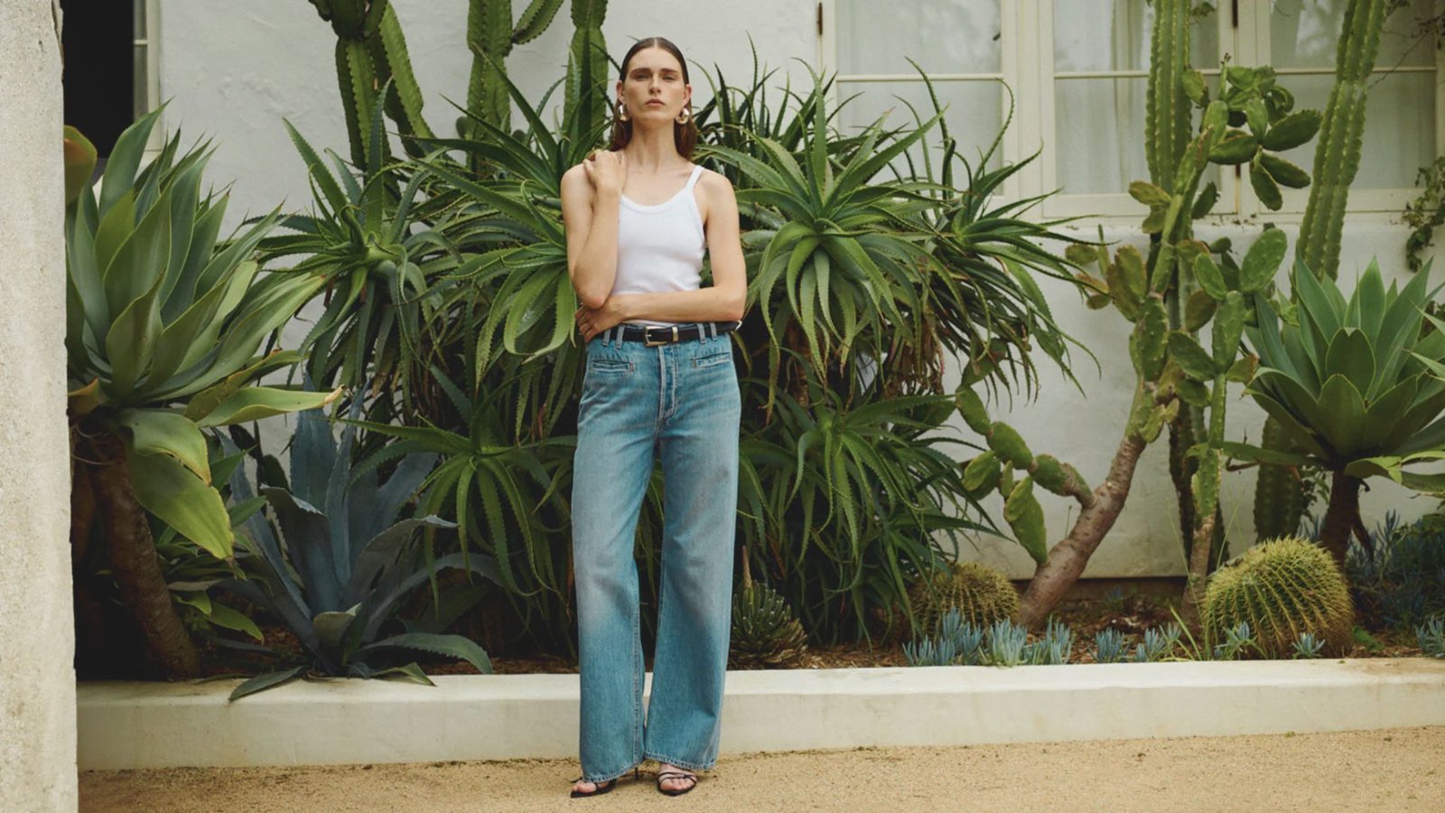 A woman in a white tank top and blue jeans stands in front of tall cacti against a clear blue sky.