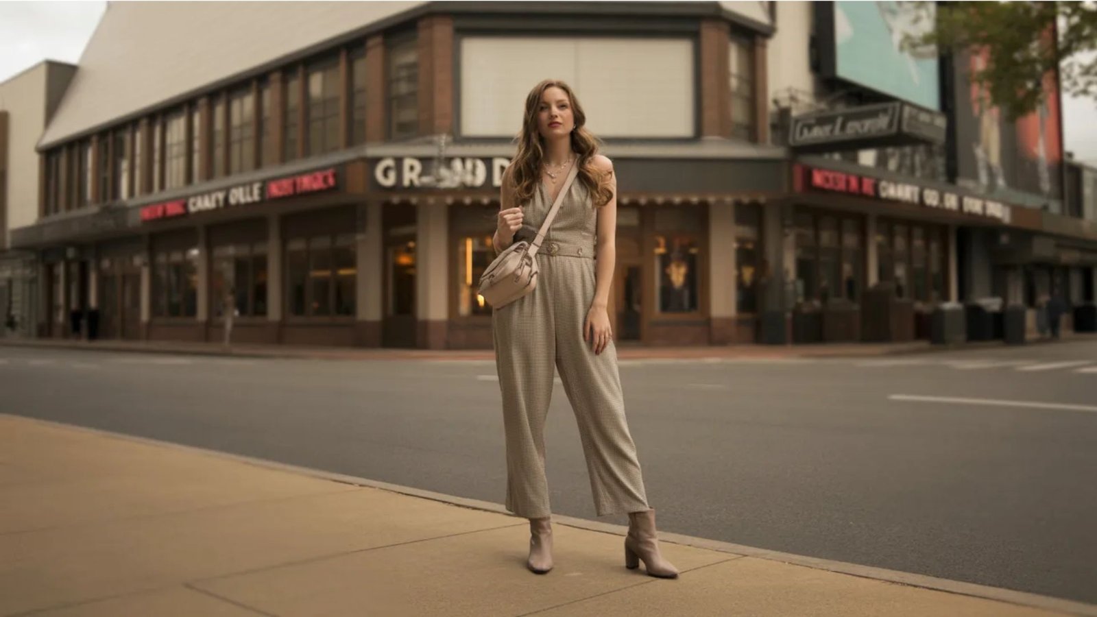 A woman in a beige jumpsuit stands confidently on the sidewalk, surrounded by urban scenery.