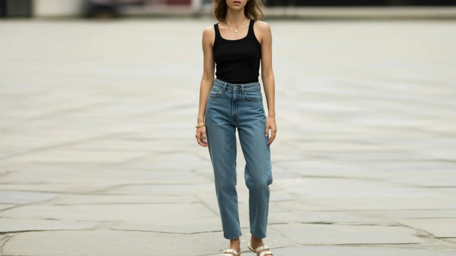 A woman wearing jeans and a black top stands confidently on a city street, surrounded by urban scenery.