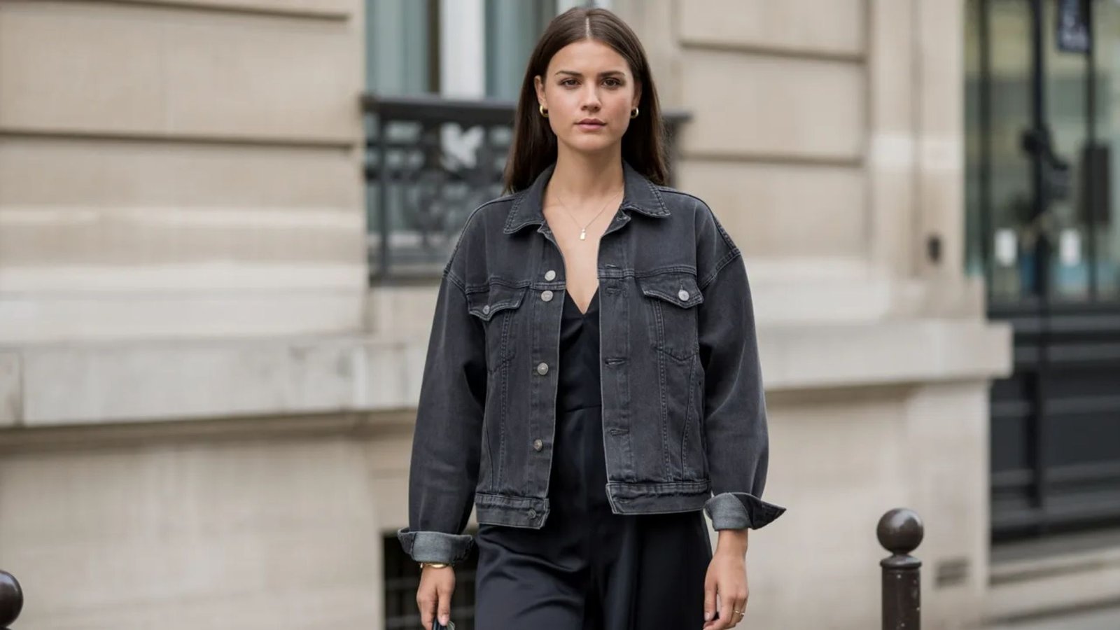 A woman wearing a black jumpsuit and a denim jacket stands confidently, showcasing a stylish and casual outfit.