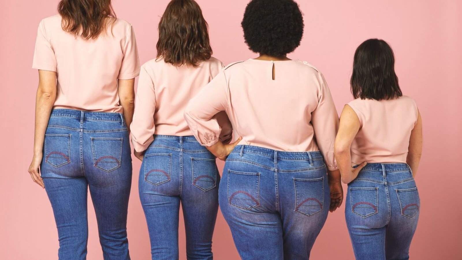  Five women in jeans pose together against a vibrant pink background, showcasing a casual and stylish look.