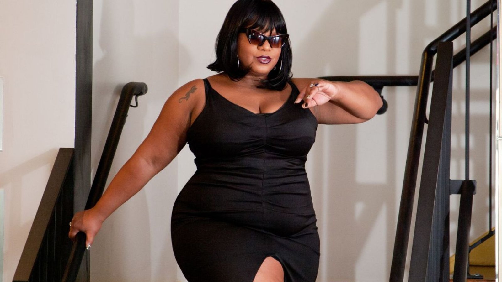 A woman wearing a black dress and sunglasses stands confidently, exuding elegance and style.