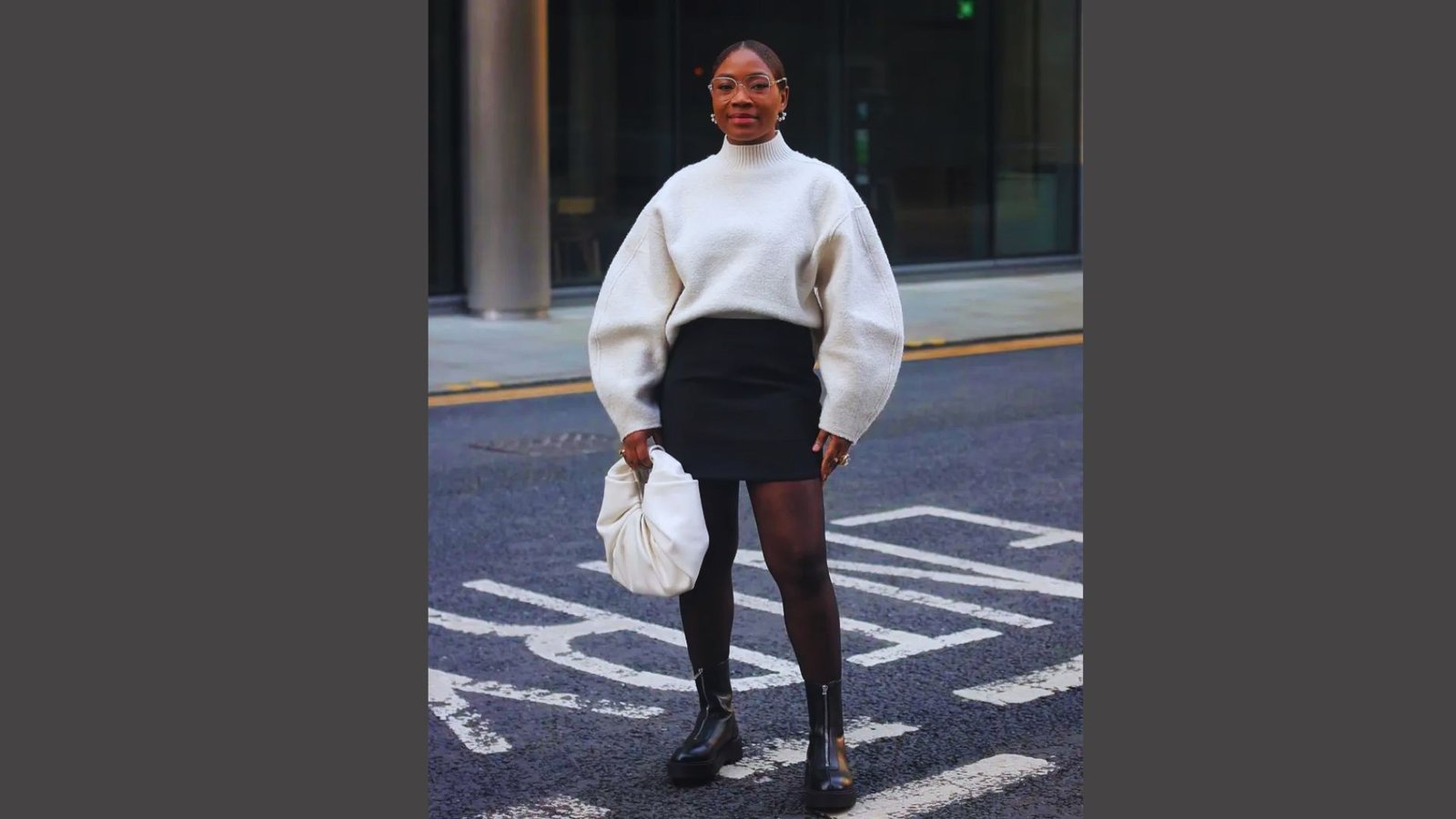 A woman wearing a white sweater and a black skirt stands confidently, showcasing a stylish and modern outfit.