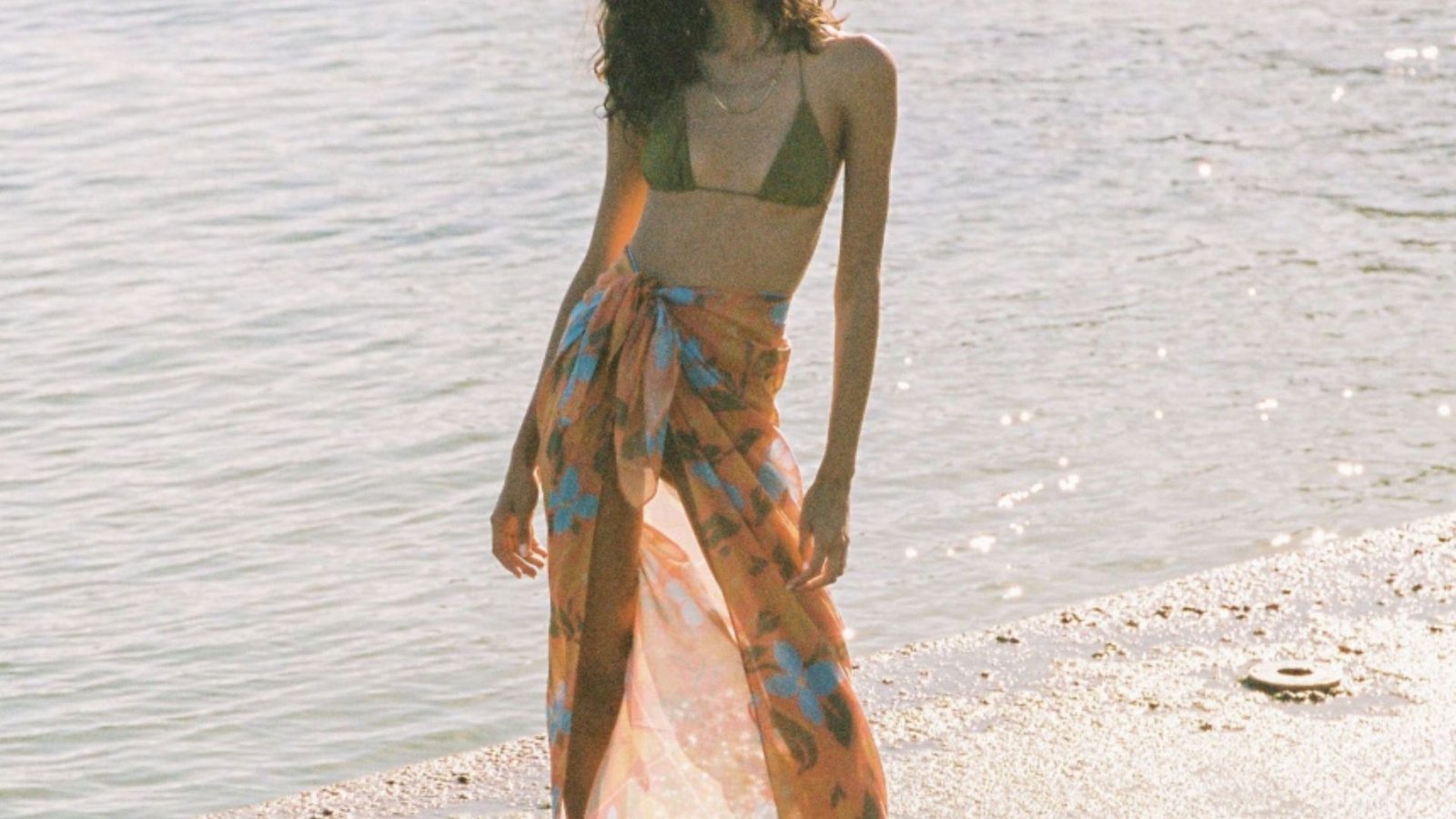 . A woman in a bikini and skirt stands on the beach, with waves gently lapping at the shore in the background.