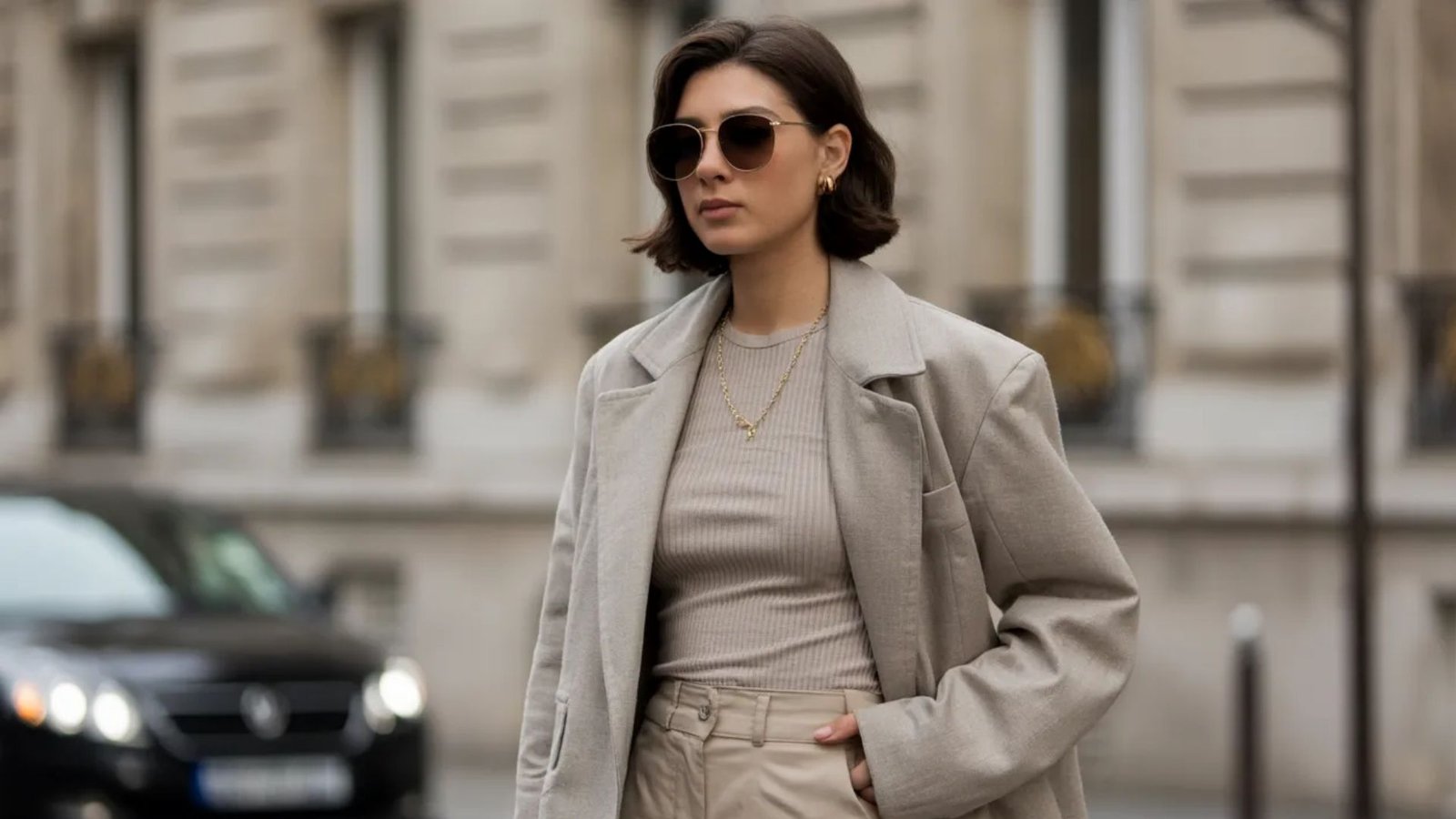 A woman wearing a beige coat and sunglasses stands confidently, showcasing her stylish outfit.