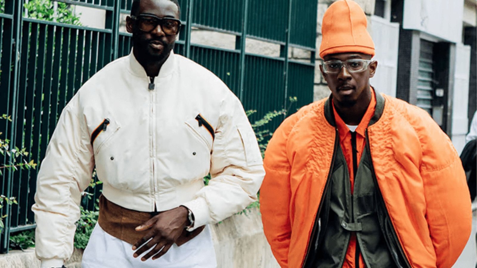 Two men in orange jackets and white pants stand together, smiling against a neutral background.