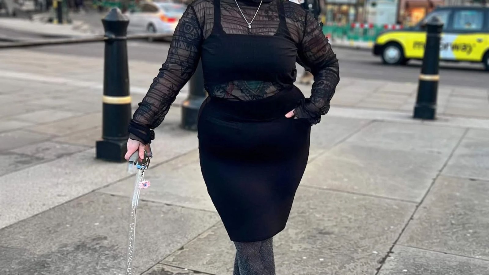 A woman in a stylish black dress and high heels stands on a sidewalk, exuding elegance.