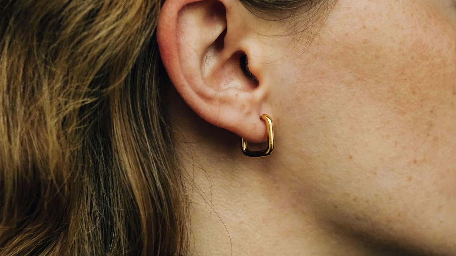 Close-up of a person's ear wearing a minimalist, gold hoop earring. The surrounding skin has freckles and light brown hair is visible, evoking a natural and simple style.