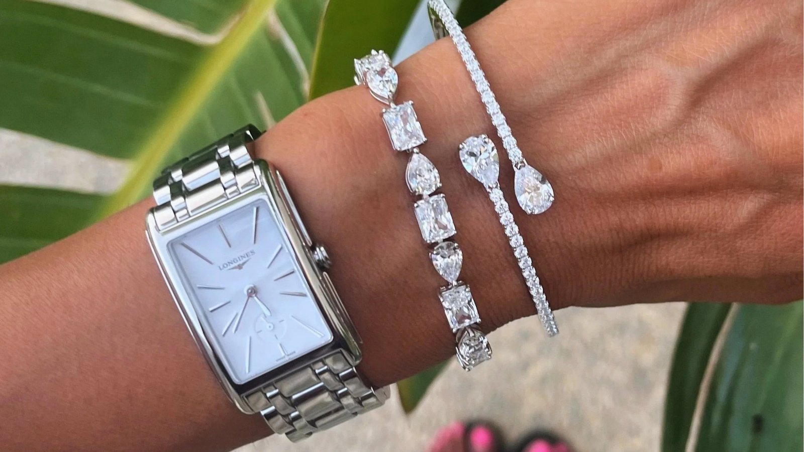 A wrist adorned with a silver watch and three diamond bracelets, featuring various cuts. The background has a green leaf, adding a tropical feel.