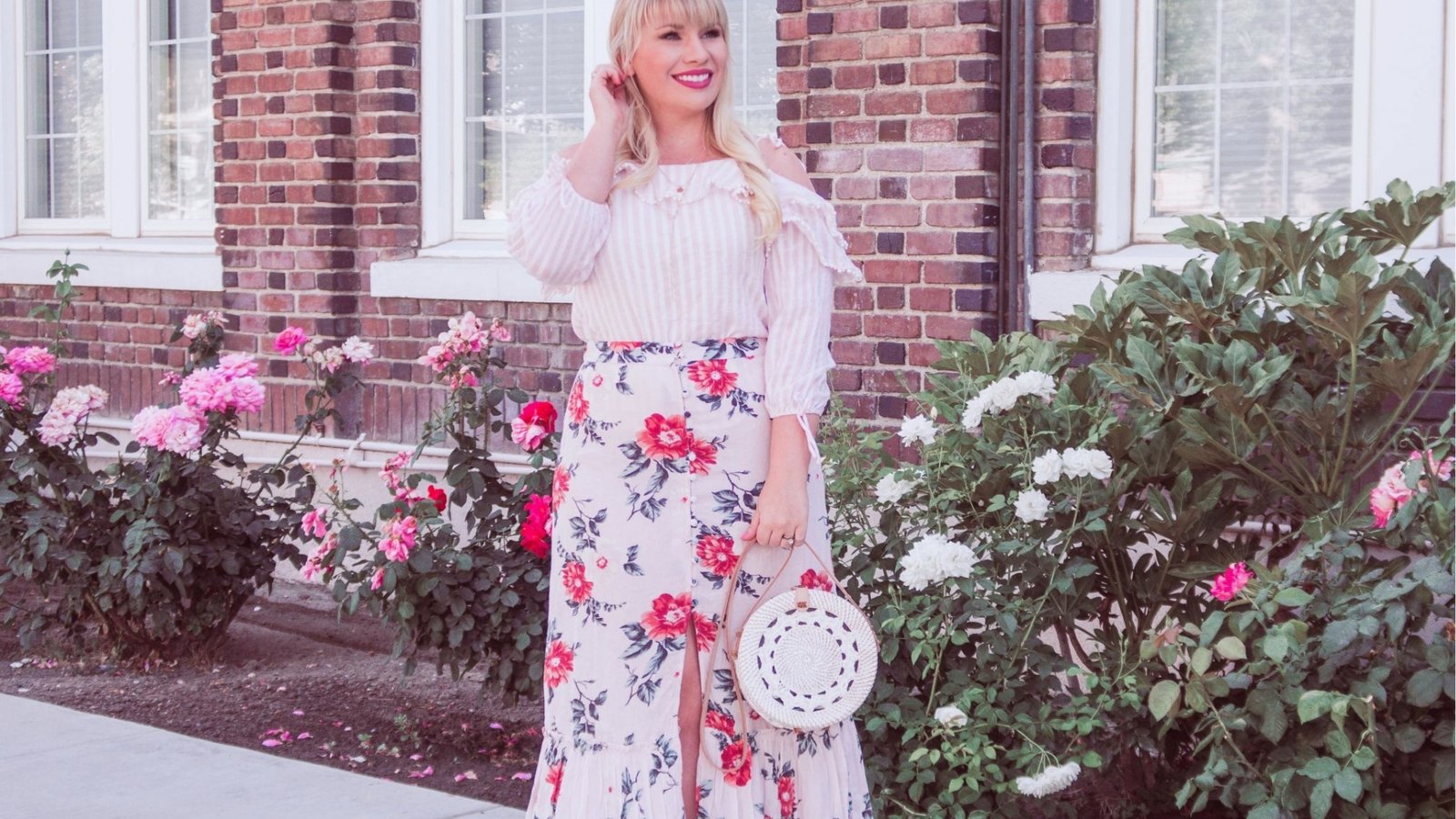 A woman in a floral skirt and striped blouse stands smiling among pink and white roses, holding a white circular bag, in front of a brick building.