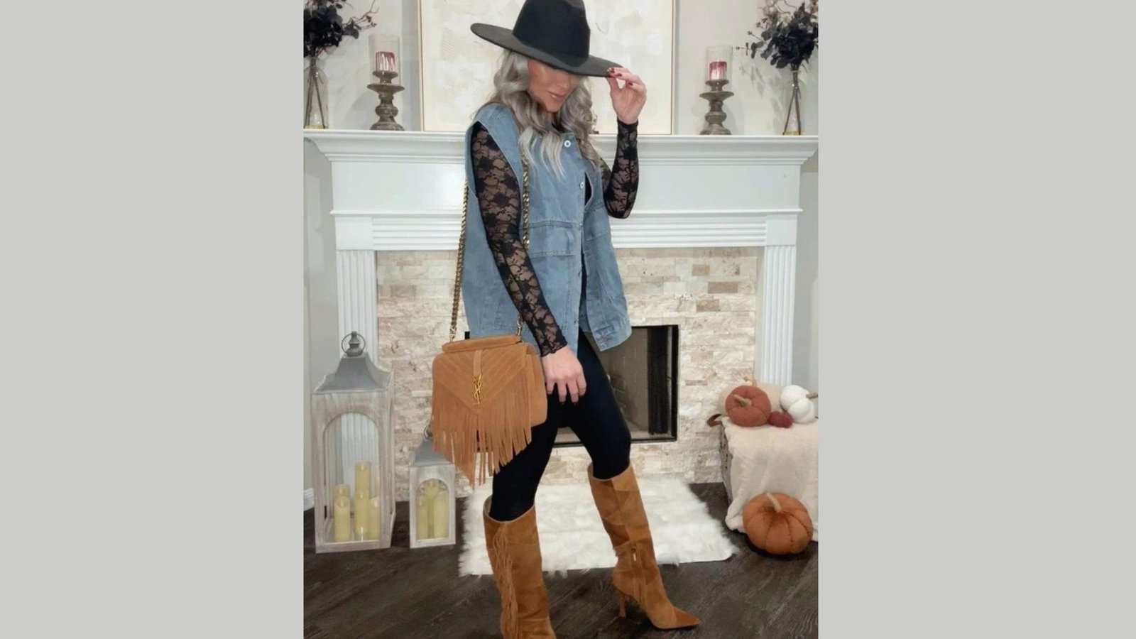A woman in a blue denim vest and black lace shirt poses by a fireplace. She wears a wide-brim hat, brown suede boots, and holds a fringed bag, creating a stylish, autumnal look.