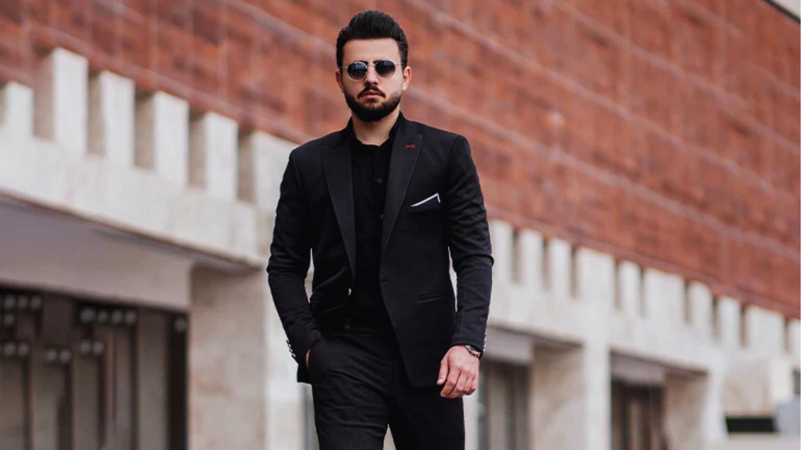 A man in a sharp black suit and sunglasses walks confidently beside a modern brick building. The scene conveys a stylish, professional tone.