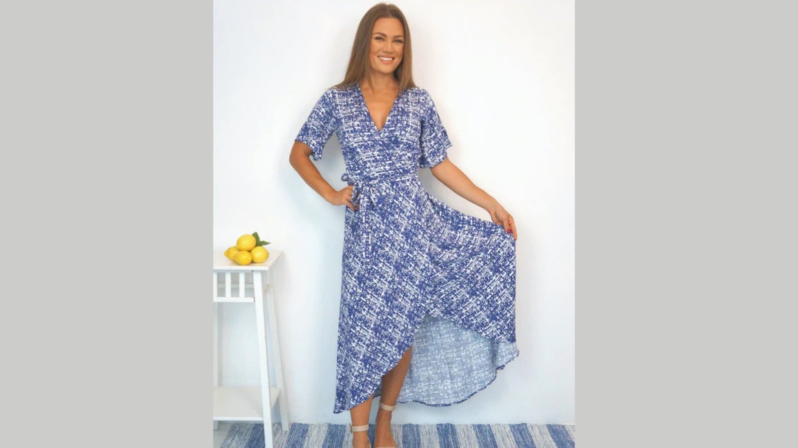Woman in a blue patterned dress poses with a smile, holding part of the dress. A small table with lemons is beside her, and the mood is cheerful and summery.