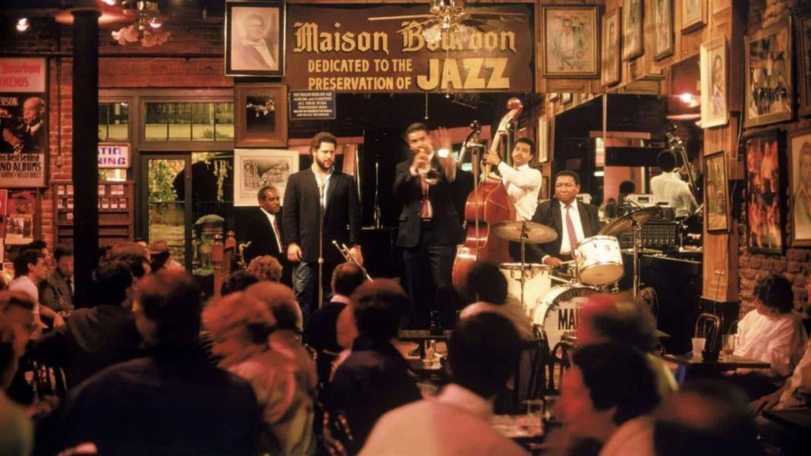 Jazz band performing at Maison Bourbon, New Orleans. A trumpeter, bassist, and drummer play energetically. Audience seated closely, creating an intimate, lively atmosphere.